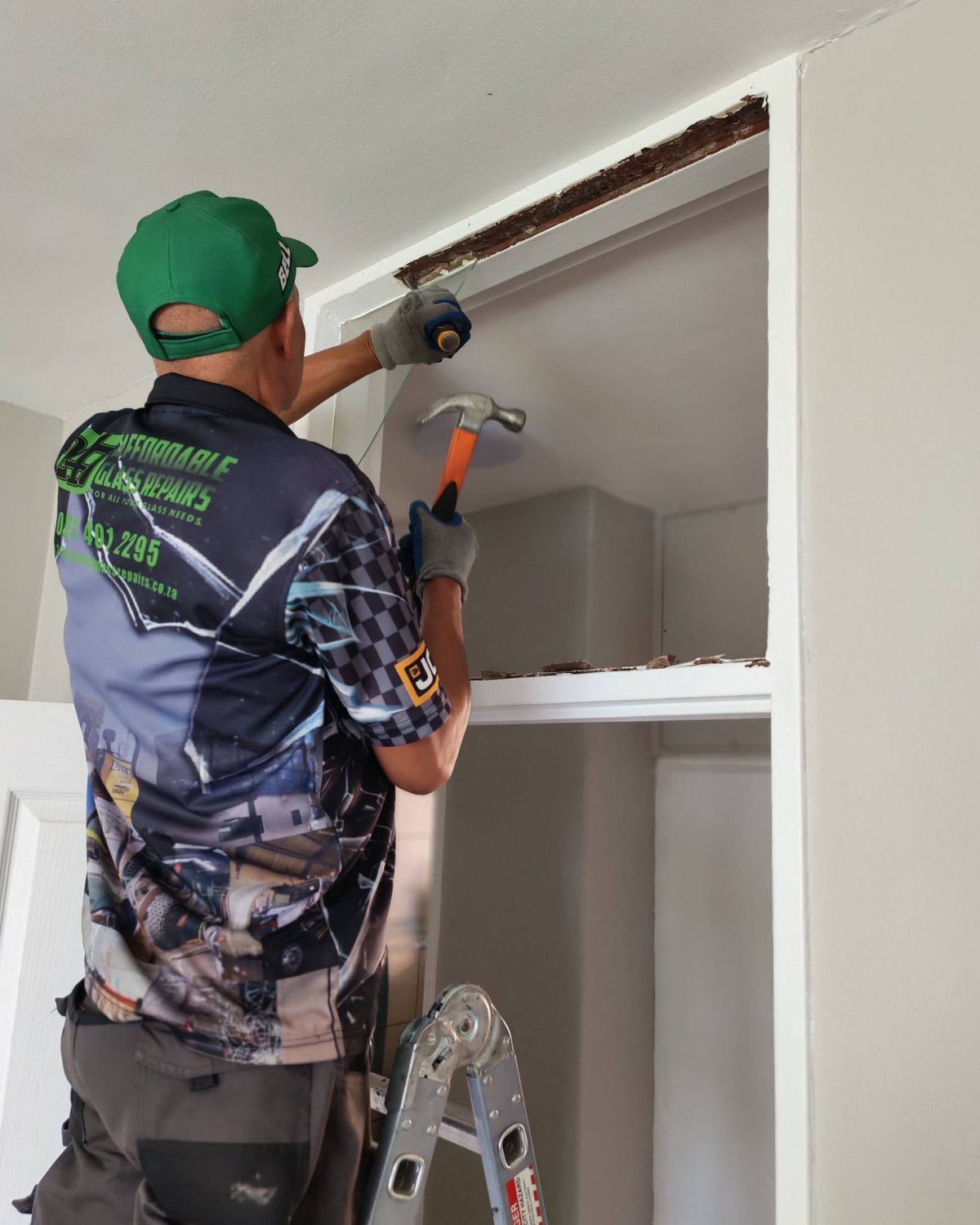 Worker in green cap installing or adjusting trim around a white door frame using a power drill and hammer.