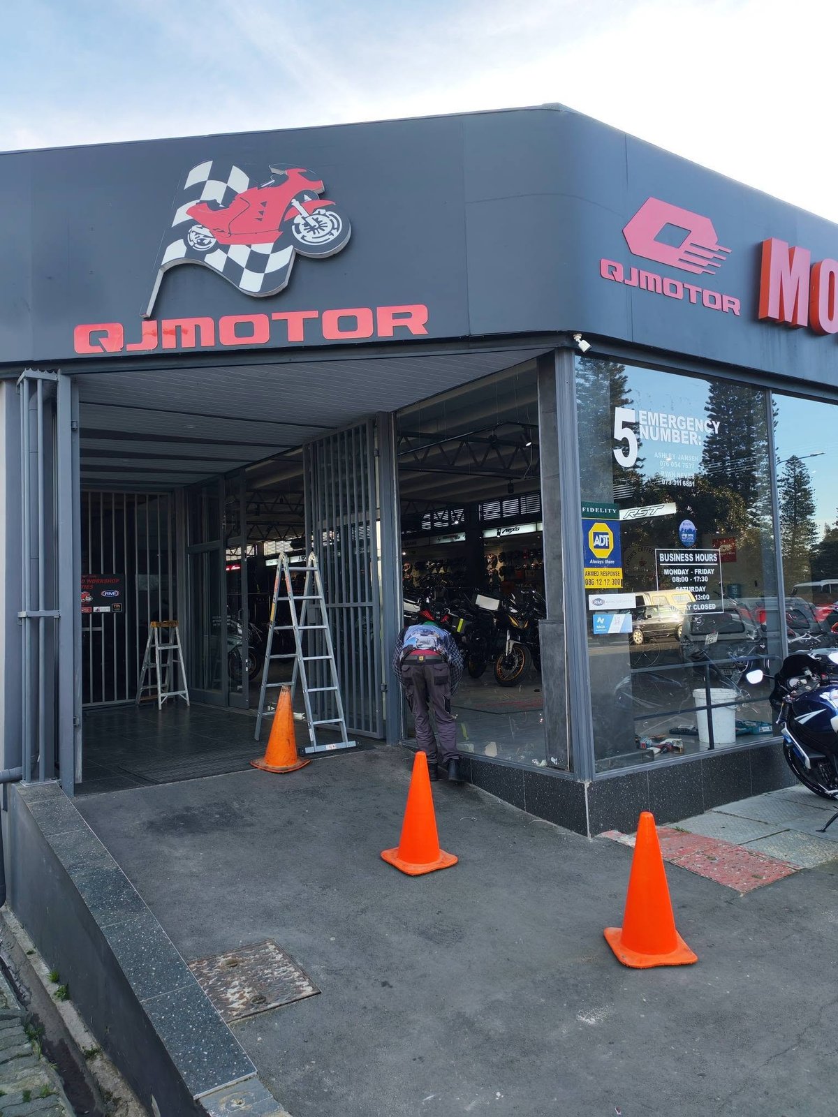 AJ Motor storefront with red motorcycle logo, metal shutters, display windows, and orange traffic cones on sidewalk while broken shopfront glass is being secured