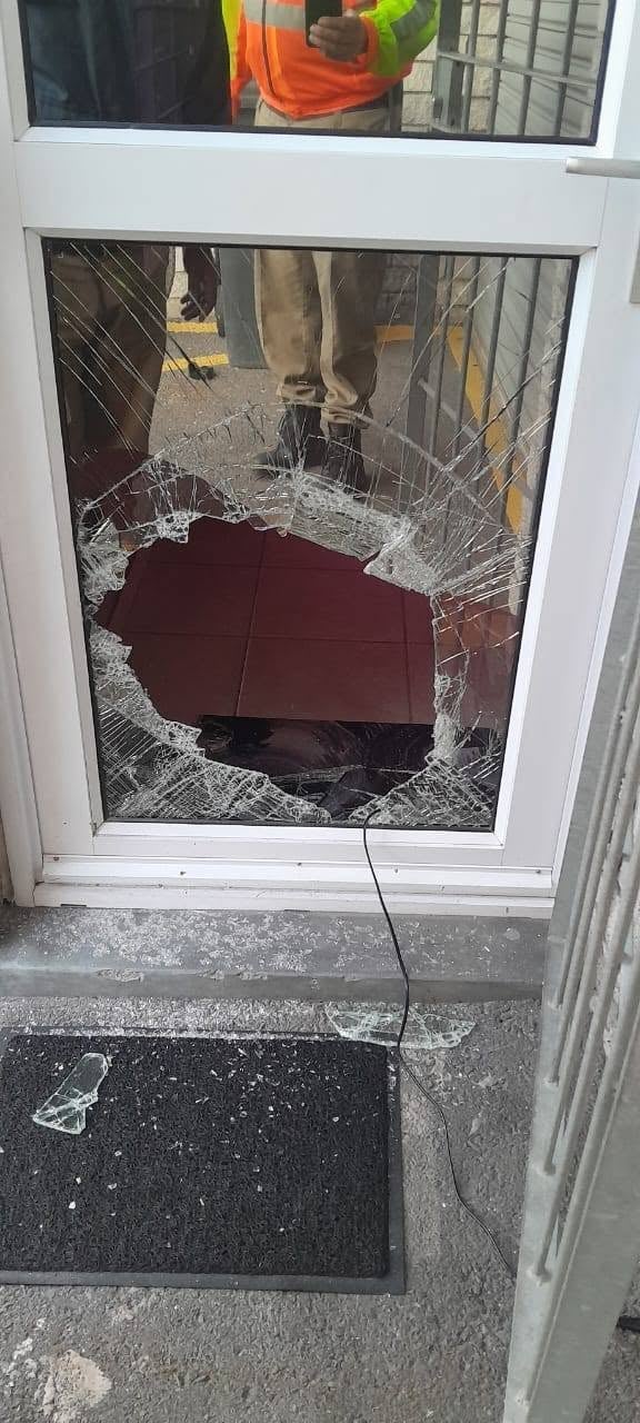 Shattered glass door with large hole covered by red tarp, workers visible outside through undamaged upper window