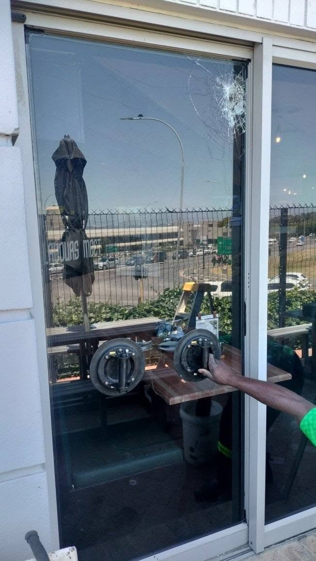 Cracked shopfront glass door at a Bellville restaurant with a spider-web impact break in the pane, while a 247 Affordable Glass Repairs technician uses glass suction lifters to lift the damaged panel out of the frame, with the restaurant's terrace seating and umbrellas behind.