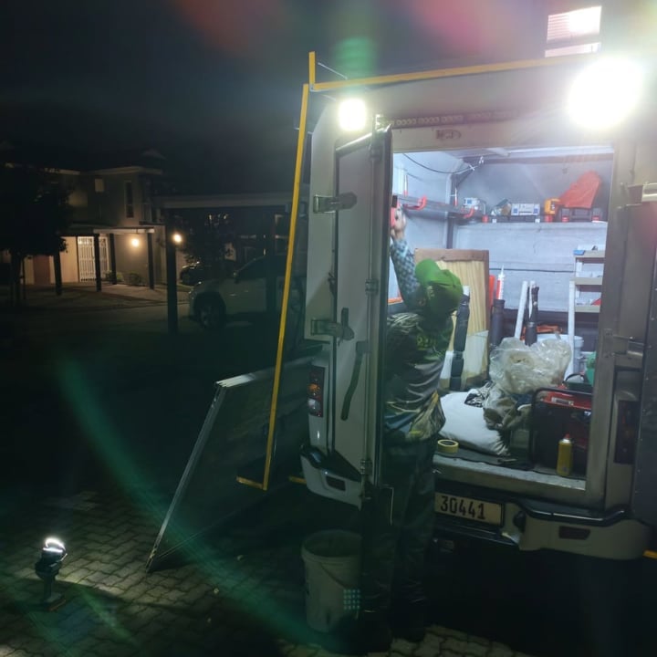 247 Affordable Glass Repairs technician in a green cap working out of the open back of the company's mobile workshop van under bright work lights at night, loading tools and glass in a Tamboerskloof residential street after dark.