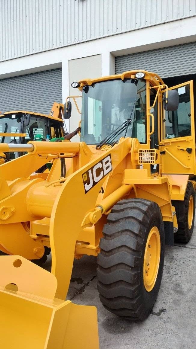 Same yellow JCB wheel loader parked in the same position, now with the new curved front windscreen fully fitted into the cab, the cab door open and the operator's seat visible through the new glass.
