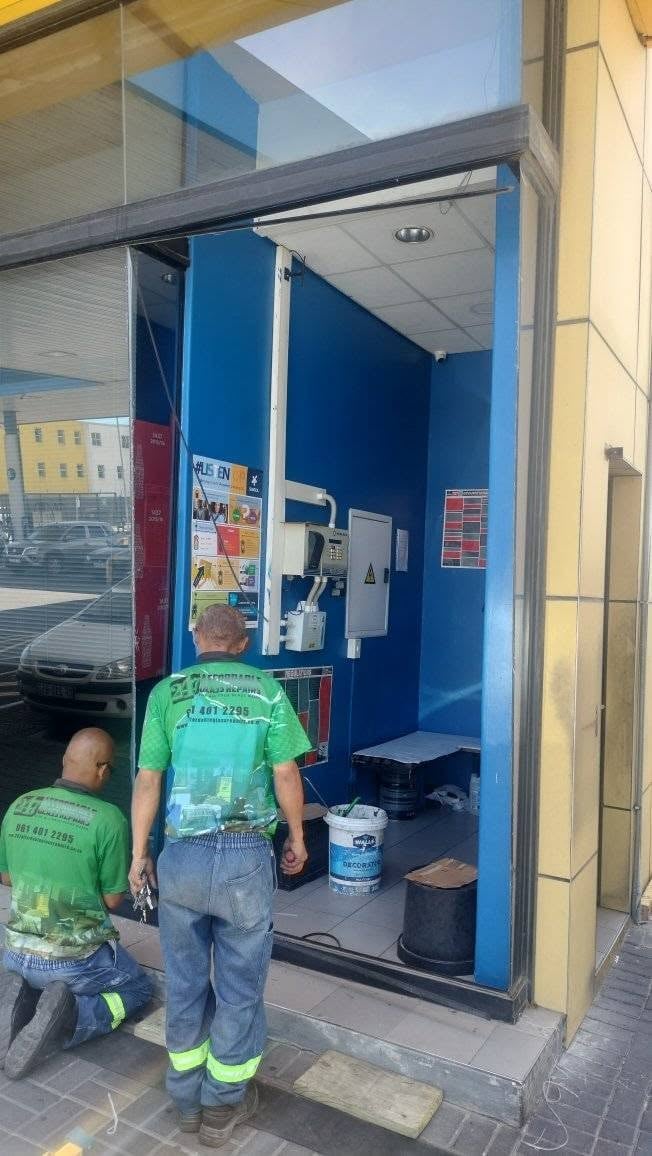 Two workers in green safety vests entering a blue-walled storefront with various supplies and equipment inside during a shopfront glass repair