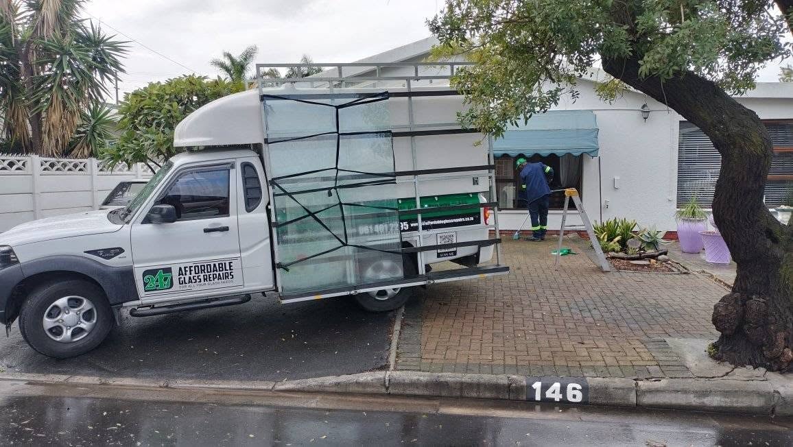247 Affordable Glass Repairs mobile glass unit parked on a brick driveway outside a Bellville home, the branded bakkie loaded with a rack of glass panels and the company's name, cell number and email on the door, while a technician in green works on a window in the background.
