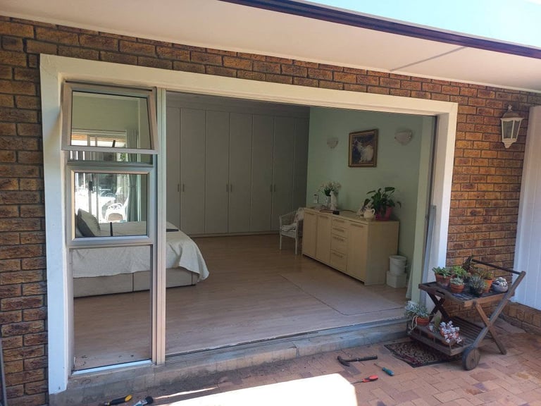 Older Oranjezicht brick home with a sliding glass door being removed — the new white aluminium frame and glass are being fitted into the brick opening, with tools on the patio and the bedroom visible through the open door.