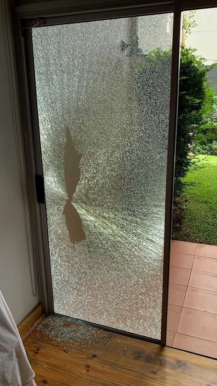Sliding glass door shattered into a spider-web pattern with glass fragments scattered across the wooden floor below and a garden visible through the broken panes