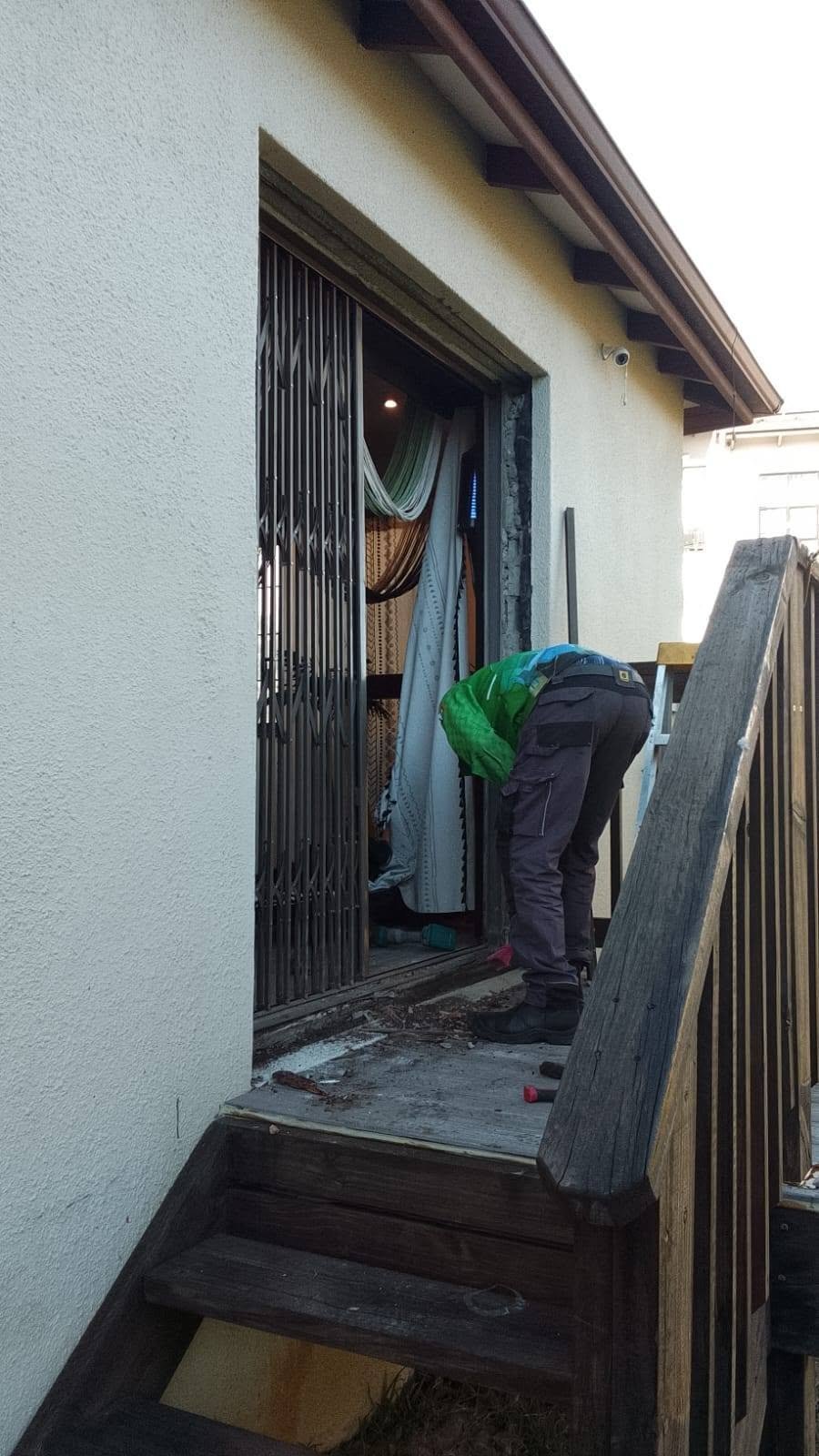 Person in green helmet bending down entering small wooden shack with black door and weathered wooden steps