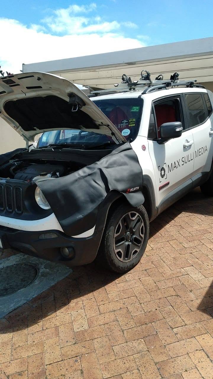 White Jeep Renegade with open hood and crumpled front end damage, parked on brick pavement under industrial roof structure