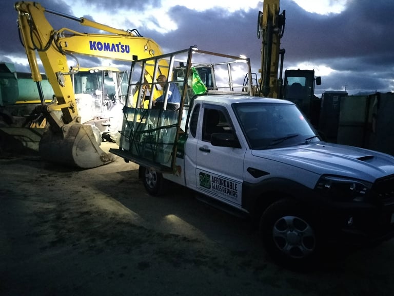 After-hours emergency earthmoving glass replacement — 247 Affordable Glass Repairs vehicle loaded with replacement cab glass alongside a yellow Komatsu excavator at an industrial site under cloudy skies