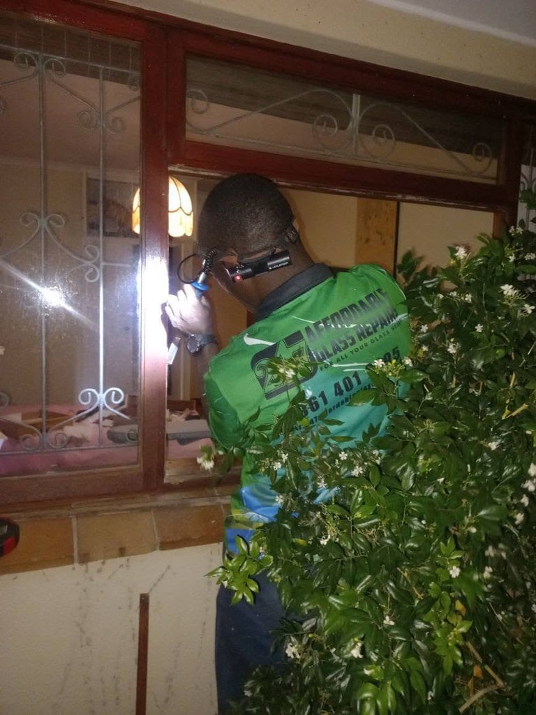 247 Affordable Glass Repairs technician in full branded green uniform with a head-mounted LED headlamp and handheld torch, working on an ornate wooden residential window with decorative grilles and leaded glass panels at night in a Devil's Peak home, with garden shrubs alongside.