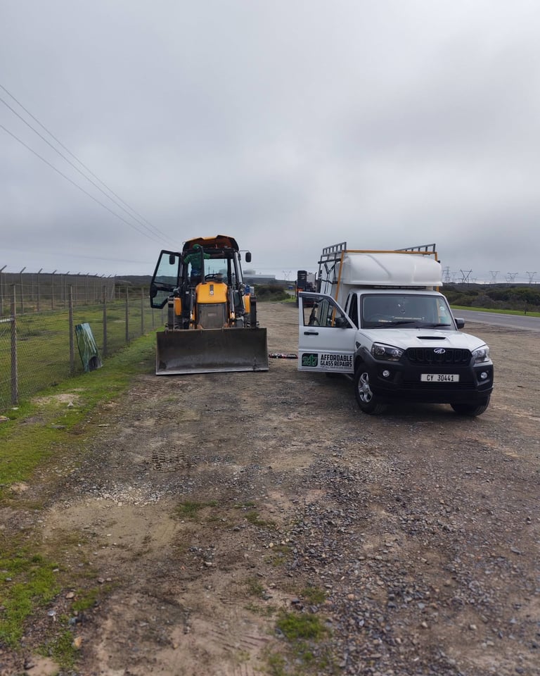 Yellow bulldozer and white 247 Affordable Glass Repairs service pickup truck with trailer parked on a gravel road beside a fenced pasture under a cloudy sky, performing an onsite earthmoving glass repair.