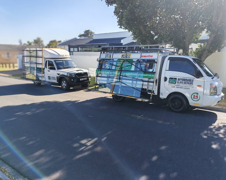 Two 247 Affordable Glass Repairs mobile workshop vehicles — a flatbed bakkie and a panel van — parked in an Oranjezicht residential street, with the company's branding, 24 HOURS emblem and contact number 061 401 2295 visible on the doors.