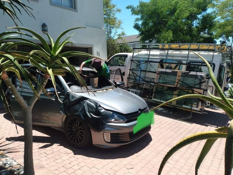 247 Affordable Glass Repairs mobile unit parked in the driveway of a Brackenfell home with its glass rack loaded, while a technician leans over the bonnet of a silver vehicle to carry out an onsite windscreen replacement, the windscreen covered in protective sheeting.