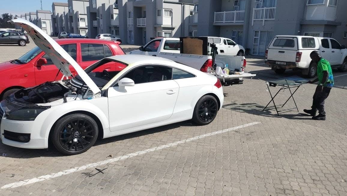 White sports car with open hood at outdoor car meet, displaying engine to attendees near other vehicles and buildings