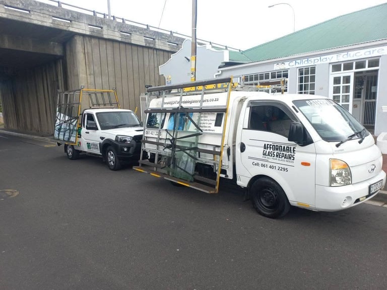 Two 247 Affordable Glass Repairs mobile workshop vehicles — a flatbed bakkie and a panel van with the company branding, cell number 061 401 2295 and service list (glass, earth moving glass, windscreens, house glass, building glass, sliding doors, mirrors, table tops and tinting) — parked on site.