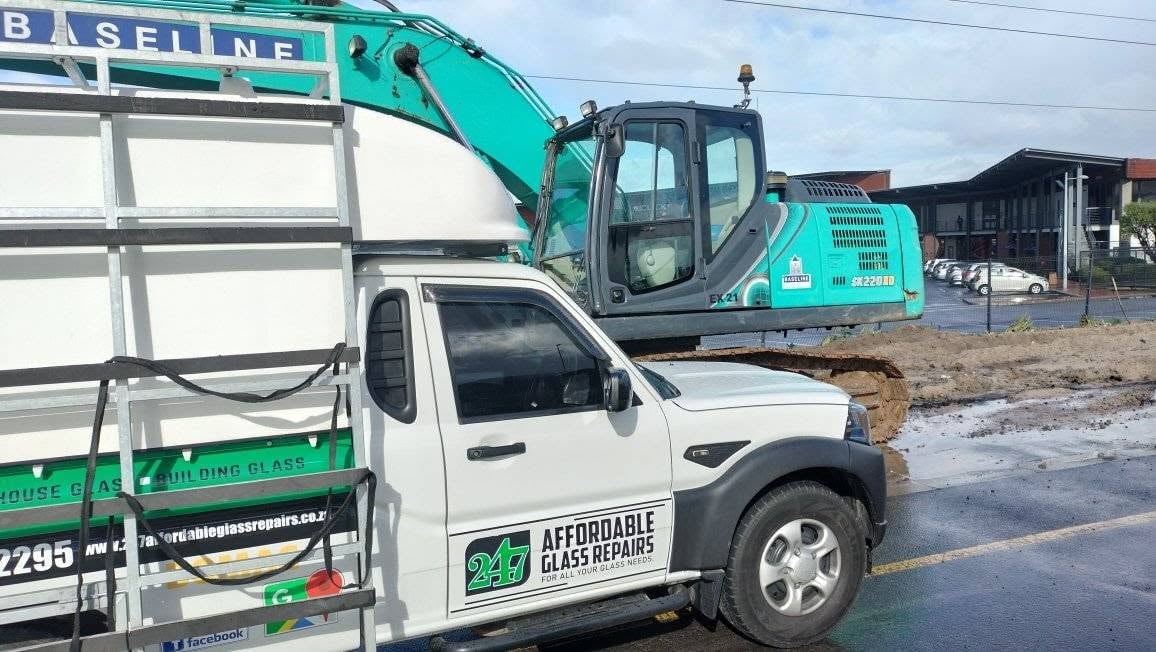 247 Affordable Glass Repairs white work bakkie with the green 247 logo and cab-mounted ladder parked next to a large turquoise Kobelco SK220 LC excavator at an industrial construction site