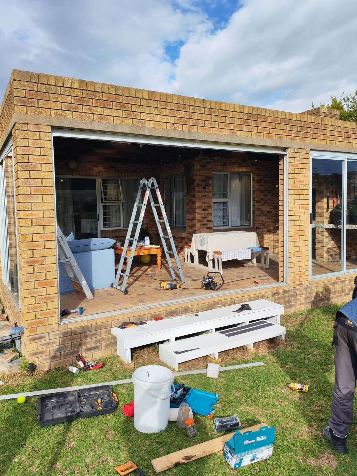 Home renovation in progress showing brick extension with open frame, ladder, tools, and materials scattered on lawn
