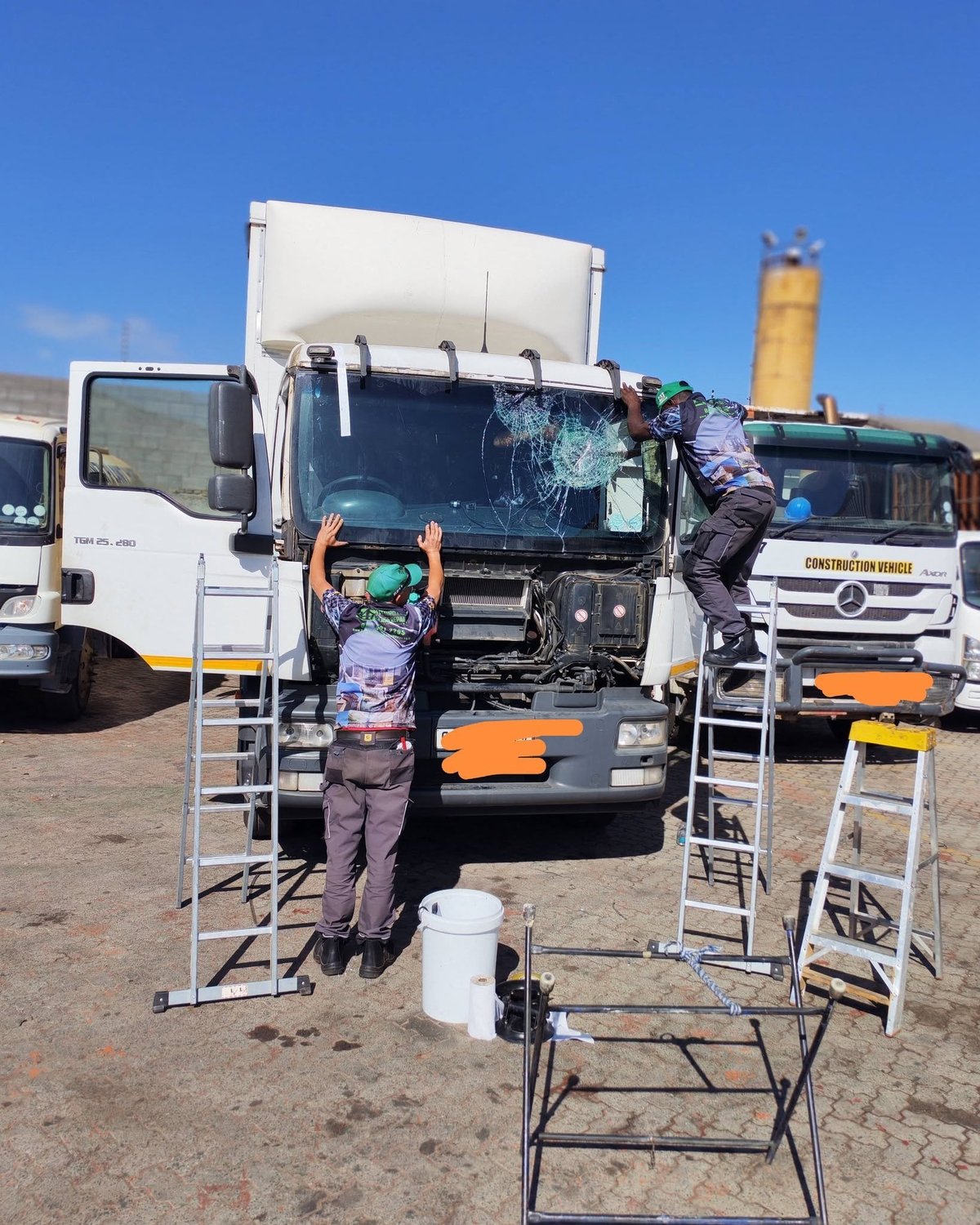 Workers performing maintenance on a large truck bed with ladders and equipment at an outdoor vehicle depot under clear blue sky