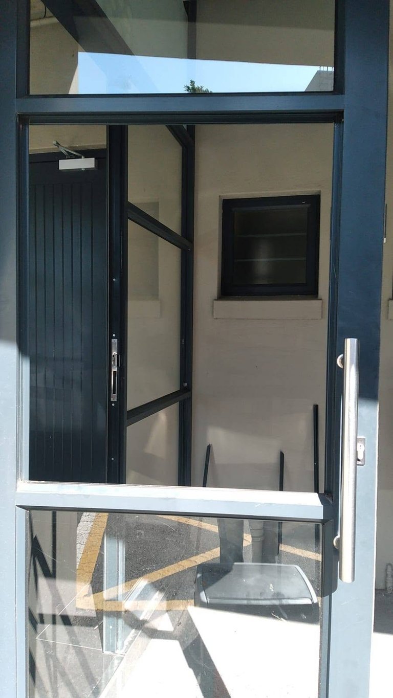Commercial glass entrance door with its damaged panel removed during a shopfront repair for a Durbanville restaurant, the dark-framed door open to show the interior hallway and kitchen area beyond, with the frame prepared ready to receive the new glass.