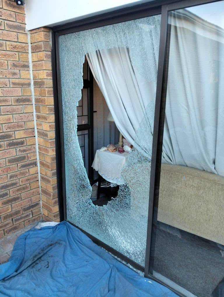 Sliding glass patio door with dark frame and a large jagged hole smashed through the centre, white curtains and a dining table visible through the gap and a blue tarp on the brick patio below.