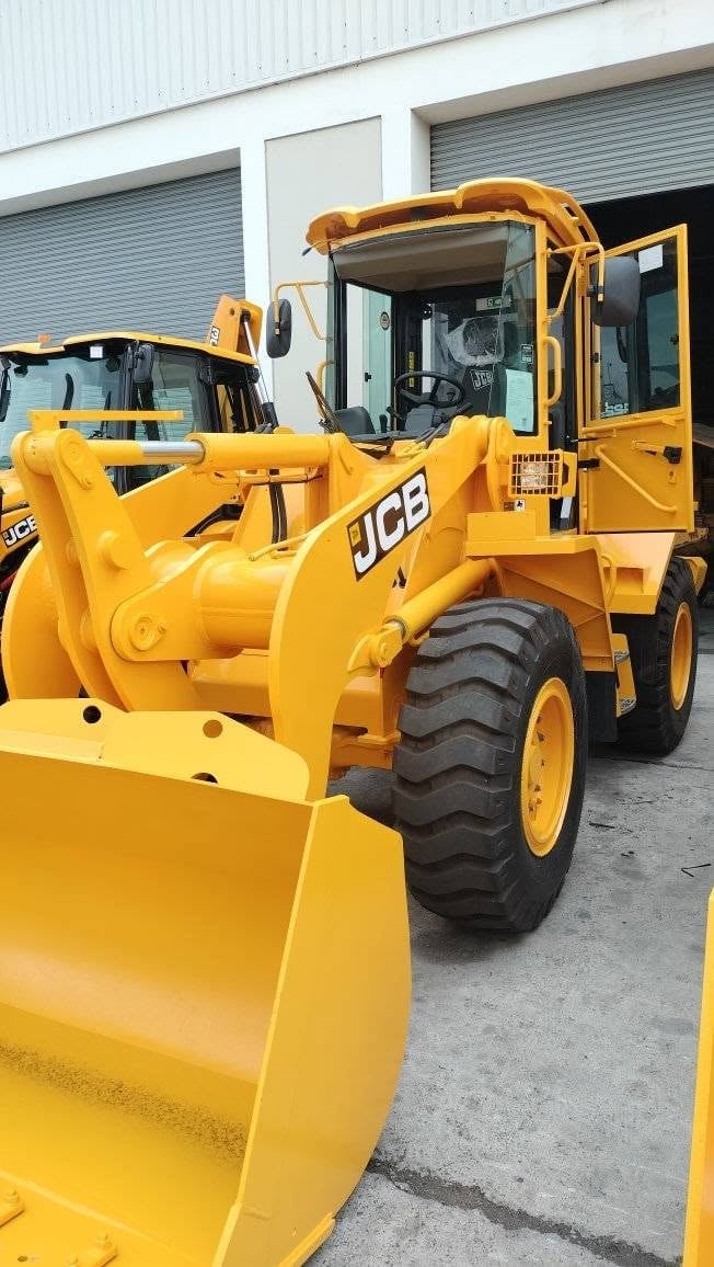 Yellow JCB wheel loader parked outside an industrial warehouse — the cab is missing its front windscreen, showing the bare black cab frame and open roof where the glass has been removed before replacement