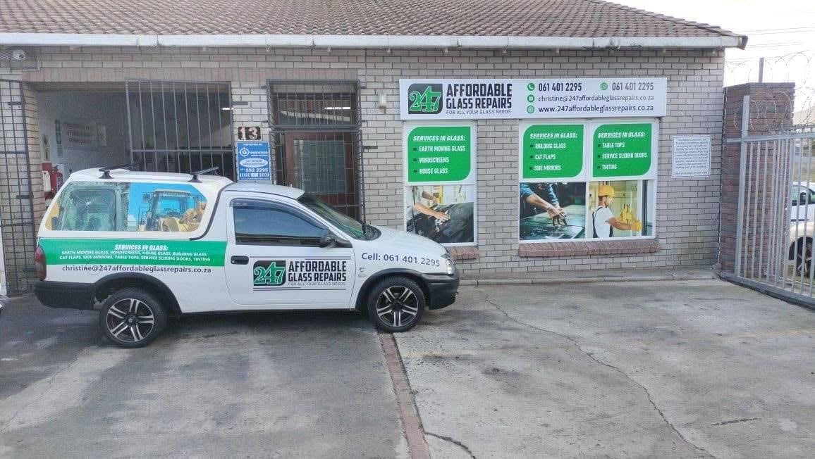 White and green 247 Affordable Glass Repairs service van parked outside the Beacon Micro Industries fitment centre, with the full-colour shop signage displaying 061 401 2295, christine@247affordableglassrepairs.co.za and the full services list: earth moving glass, windscreens, house glass, building glass, cat flaps, side mirrors, table tops, service sliding doors and tinting.