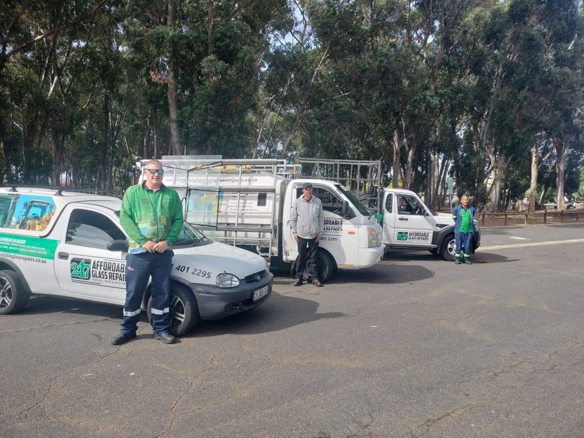 The 247 Affordable Glass Repairs team — three staff in green high-visibility shirts standing with the company's three white-and-green mobile workshop vehicles parked in a tree-lined Cape Town street on a sunny day.