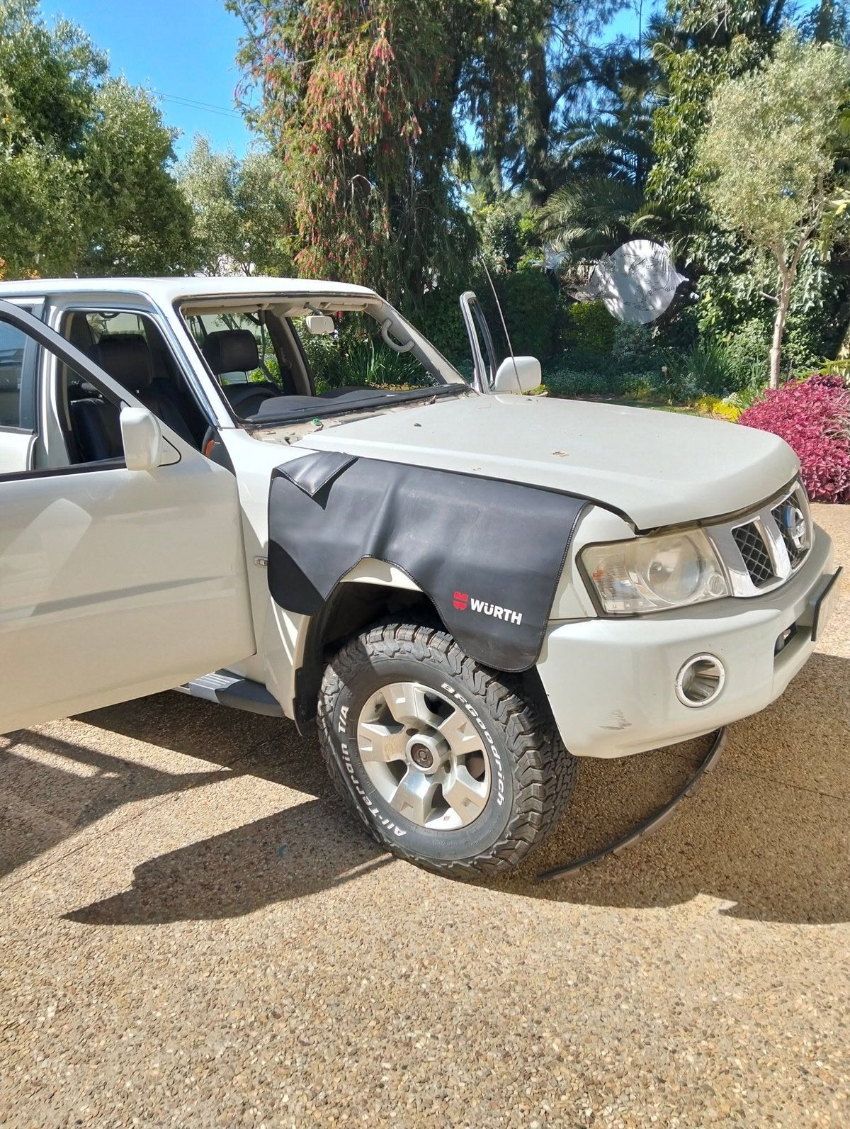 White pickup truck with driver door open and black engine cover displayed, parked in driveway with trees and flowering shrubs in background