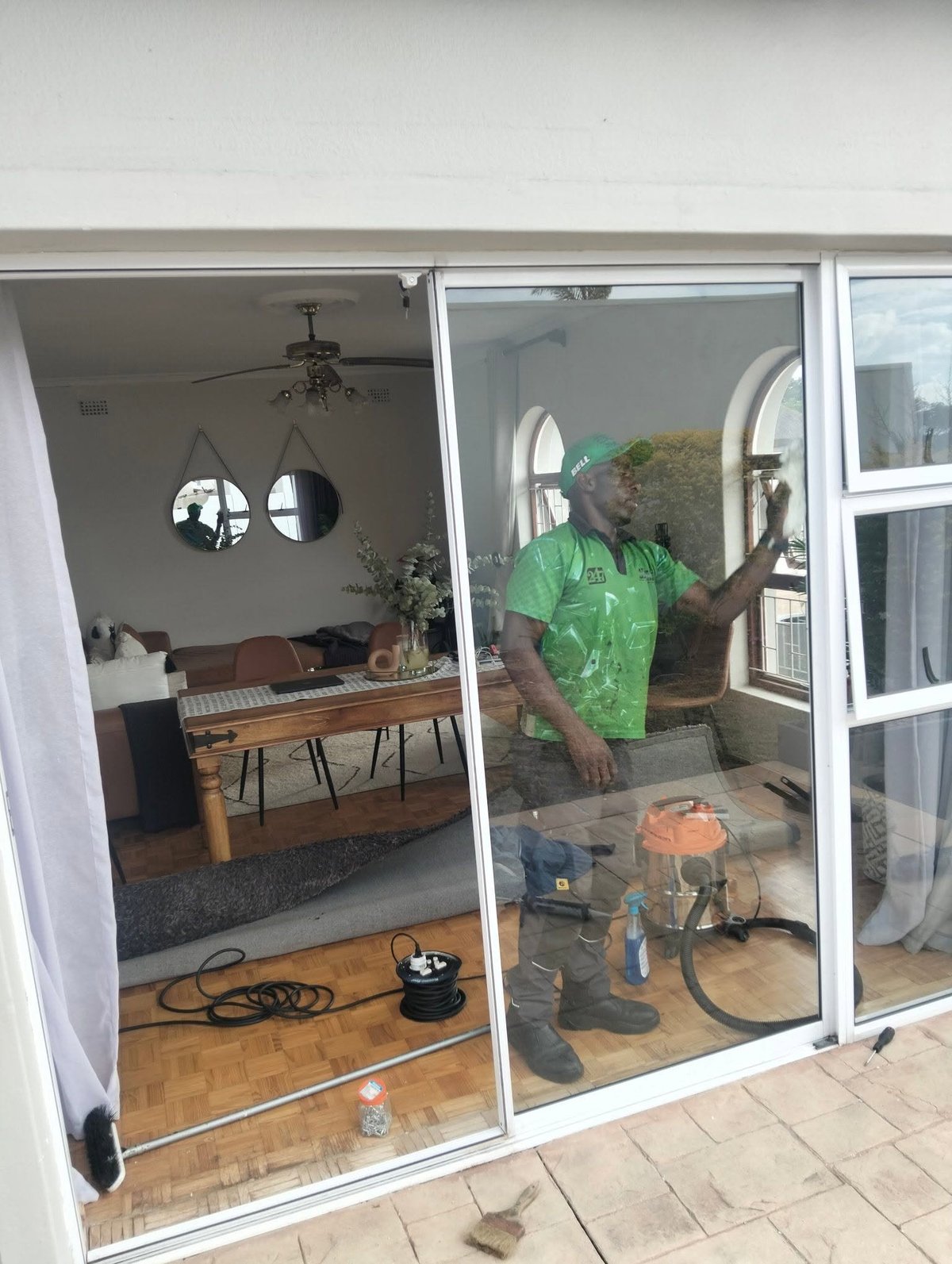247 Affordable Glass Repairs technician in a green shirt and cap refitting the inside of a newly installed sliding glass door, with an industrial vacuum and tools on the parquet floor