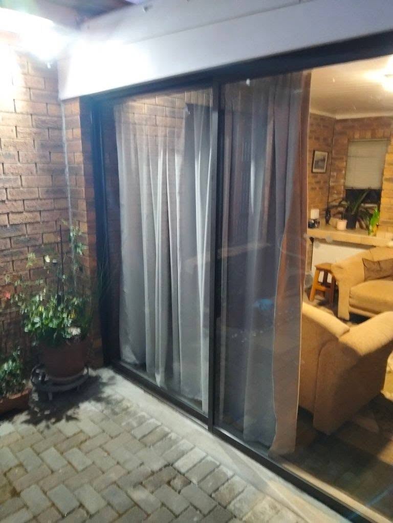 Same dark-framed sliding patio door after the smashed pane was replaced, the new clear glass reflecting the brick paving and a potted plant on the patio, with sheer curtains and the modern lounge visible inside.