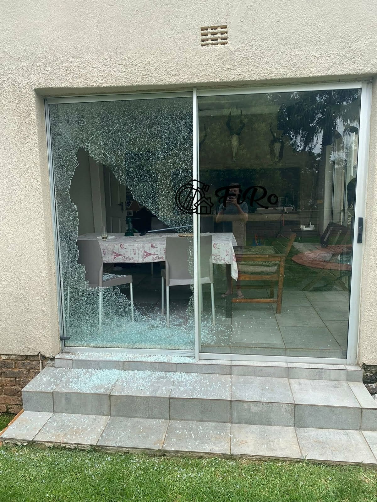 Broken sliding glass door with shattered lower pane and shards of glass scattered across the patio steps in front of a residential dining room