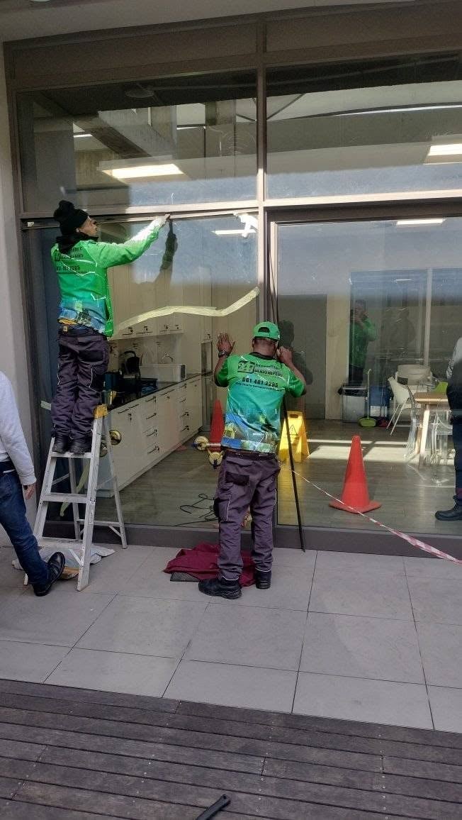 Two 247 Affordable Glass Repairs technicians in green branded work shirts fitting a large replacement glass pane into a full-height window frame on a Durbanville property, one working from a step ladder and the other steadying the glass below, with safety cones and barrier tape marking the work area.