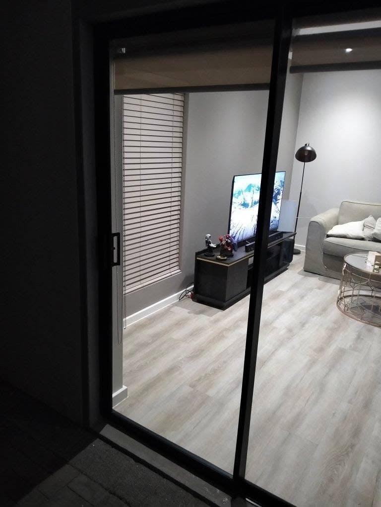 Newly installed sliding glass door in a Higgovale bedroom, with the modern bedroom (bed, TV on a black stand, floor lamp and a window with venetian blinds) visible through the clear glass.