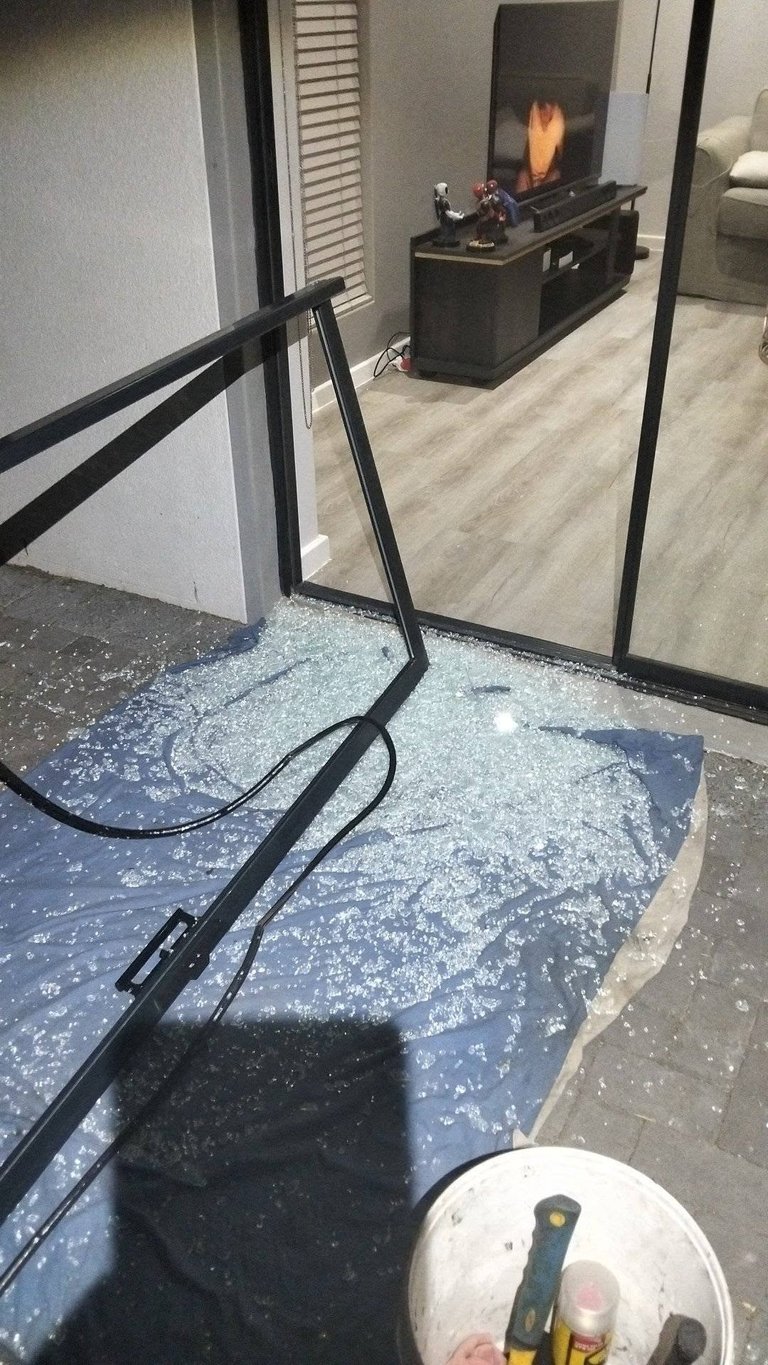 Shattered sliding door glass in a Higgovale home, with broken fragments scattered across the patio, cleaning tools in the foreground, and the modern living room with a TV and fireplace visible through the broken pane.