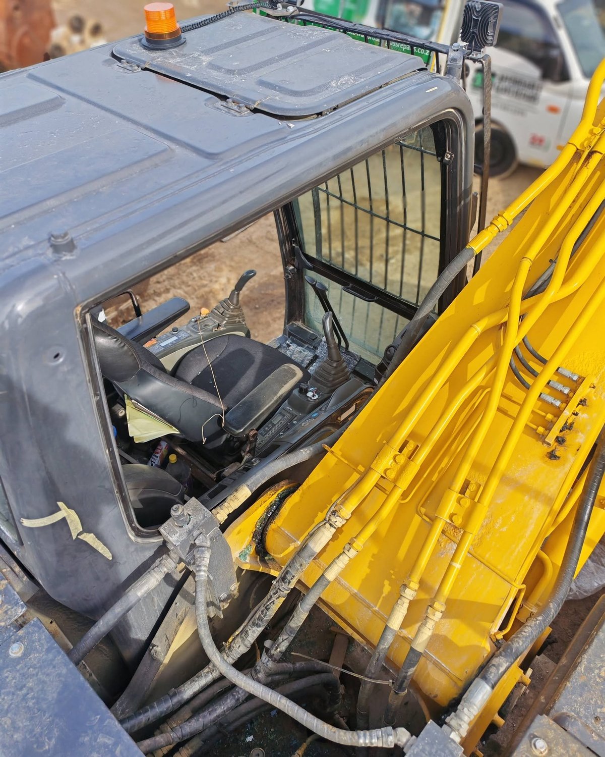 Gray utility vehicle cab with open driver's door and yellow hydraulic equipment attached to the side, awaiting glass replacement.