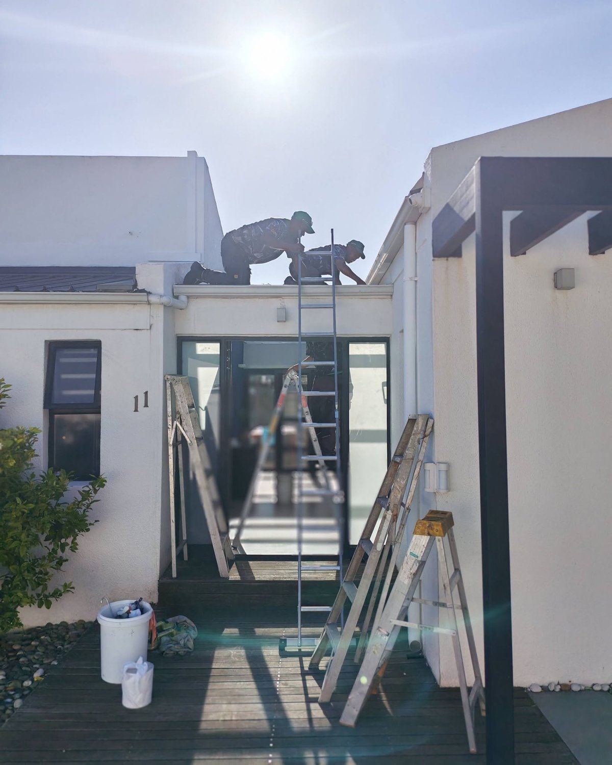 Construction site between white residential buildings with ladders, materials, and a worker on the roof frame.