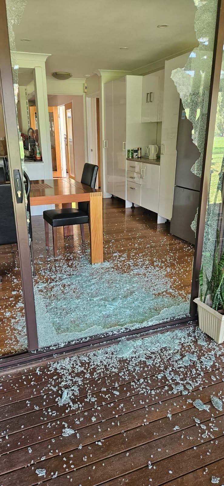Broken glass door and window scattered across kitchen entrance floor, showing damage to home interior with cabinet and appliances visible inside.