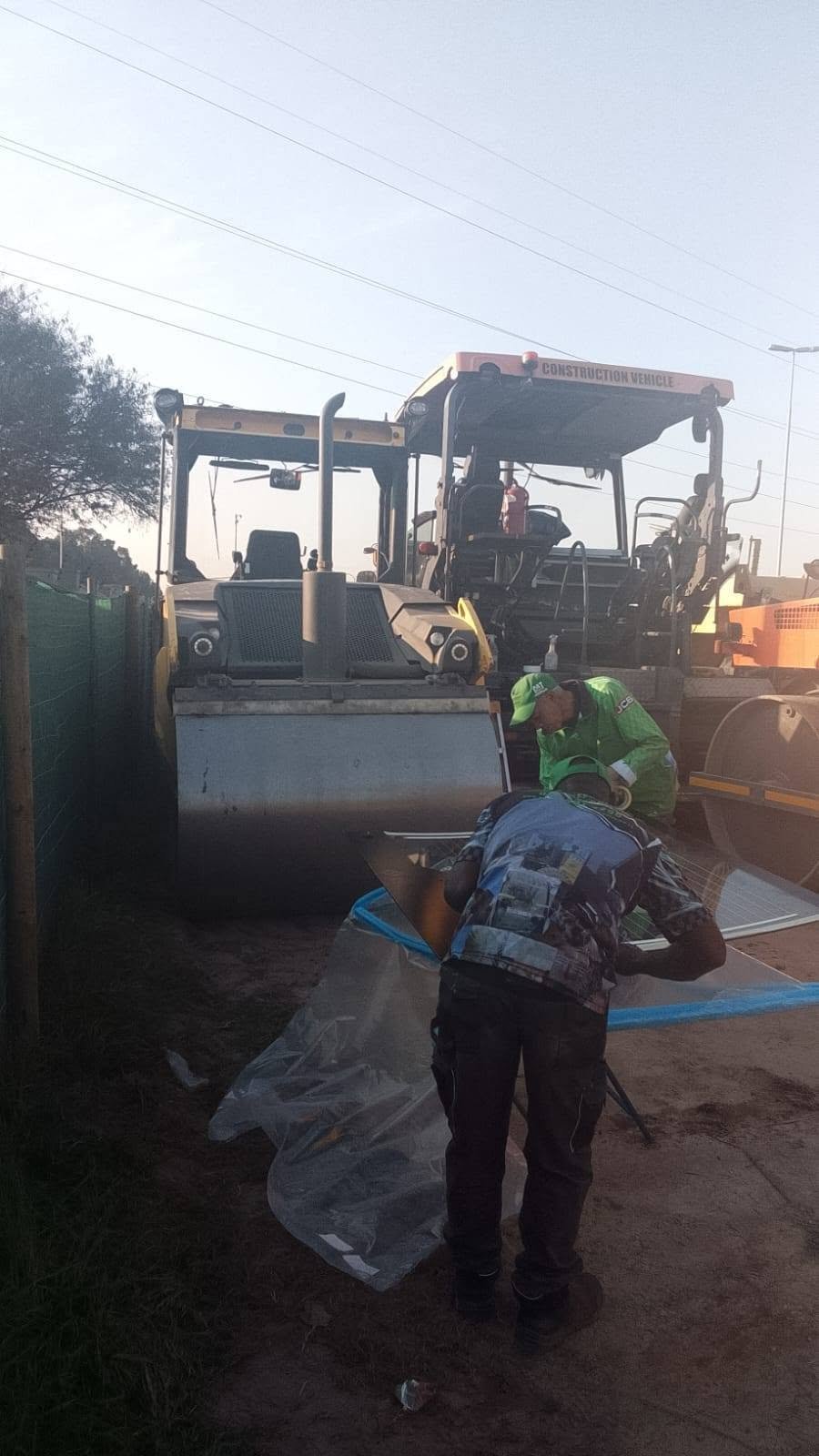 Road worker in green safety vest handling a sheet of plastic near a yellow asphalt roller compactor on a construction site.