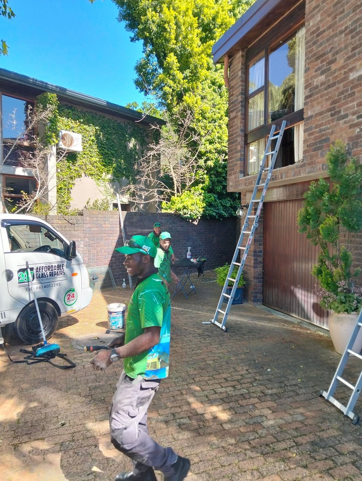 Workers in green uniforms perform maintenance on a brick building with a ladder and service van in a residential courtyard