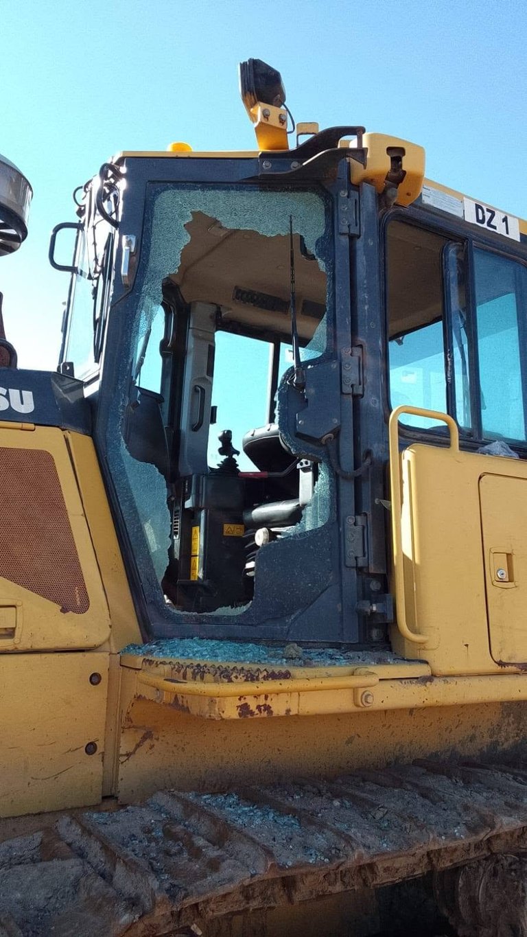 Close-up of a damaged yellow and blue Komatsu excavator cab with a shattered side window.