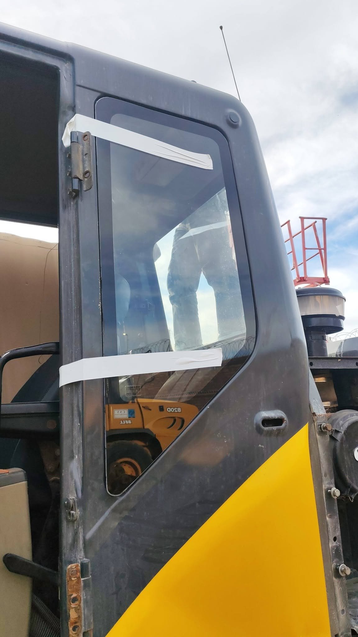 Close-up of an earthmoving machine's cab door with new glass fitted, white trim and yellow lower panel, set against a cloudy sky