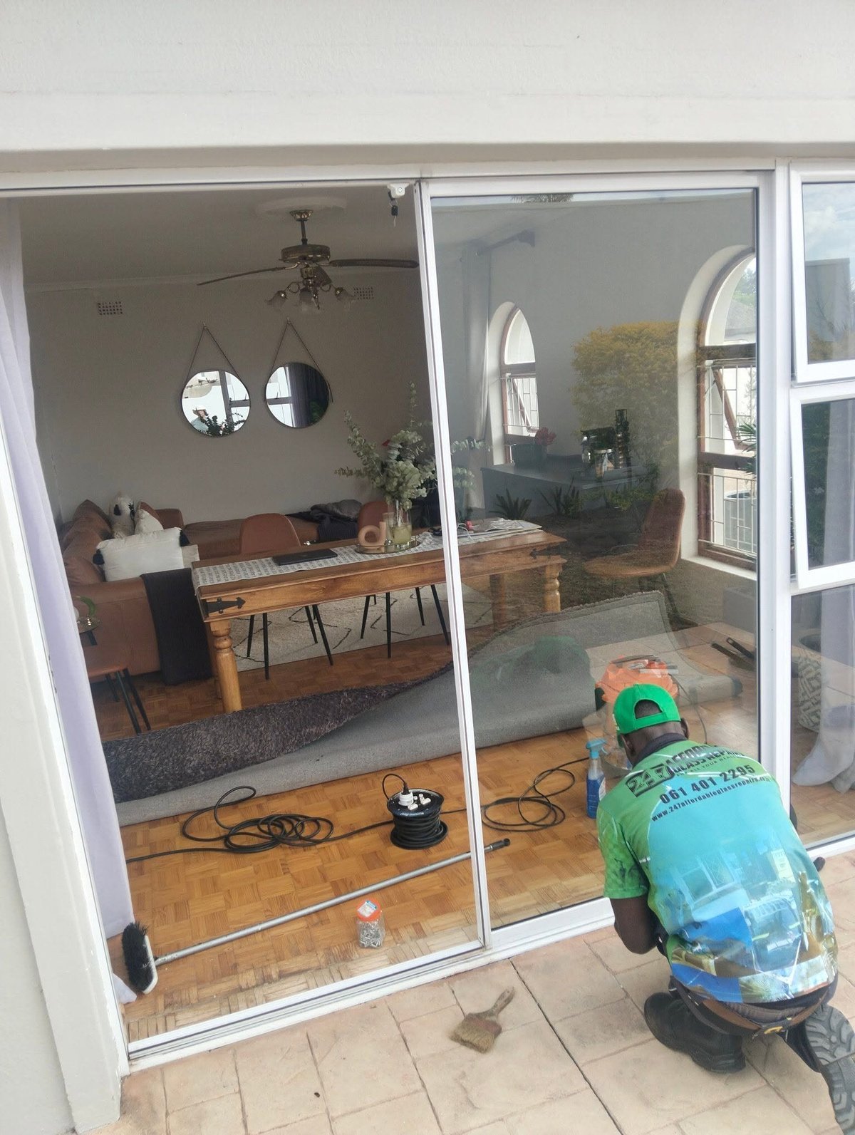 247 Affordable Glass Repairs technician crouched outside the patio, adjusting the bottom track of the newly fitted sliding glass door with the modern living room visible through the finished glass