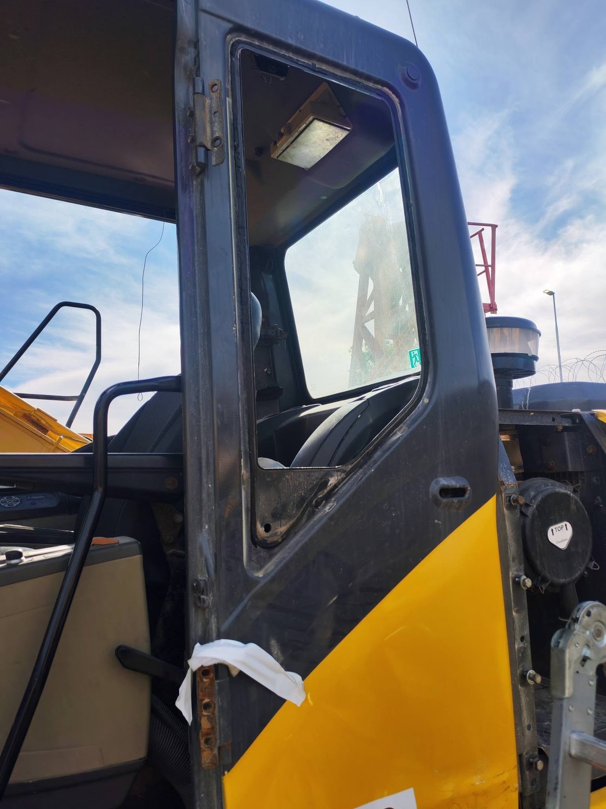 Close-up of a yellow and black construction vehicle door with an open window and industrial equipment visible in the background.