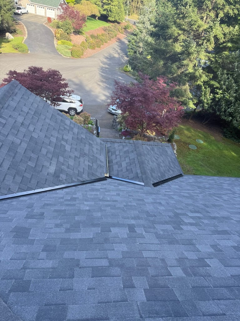 Aerial view of a residential roof with dark shingles overlooking a driveway with vehicles and landscaped yard below