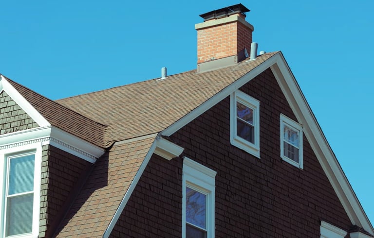 Close-up of a dark-shingle pitched roof with multiple dormer windows, brick chimney, and clear blue sky
