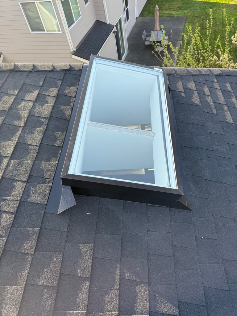 Aerial view of a white skylight with two panes installed on a dark gray asphalt shingle roof, with house exterior and yard visible