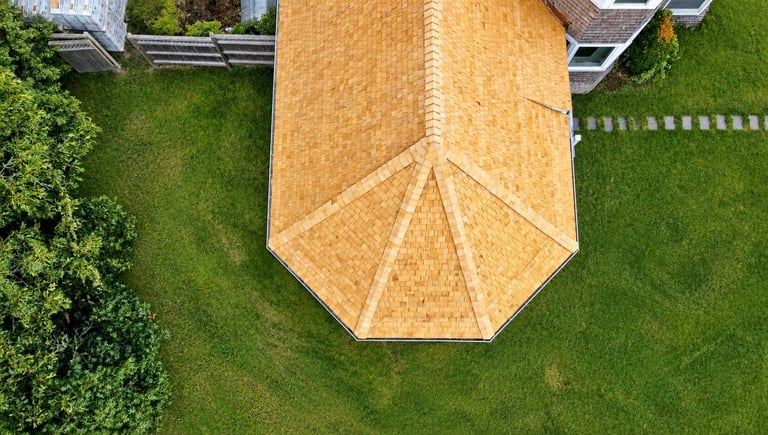 Aerial view of a house with distinctive golden-brown shingled roof sections forming a geometric pattern, surrounded by green lawn and hedges.