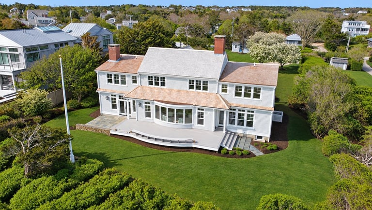 Aerial view of a large white two-story house with brick chimneys on a manicured lawn surrounded by green trees