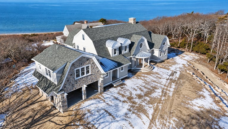 Aerial view of a large gray and white coastal home with multiple dormers overlooking the ocean, surrounded by bare trees and patchy snow.