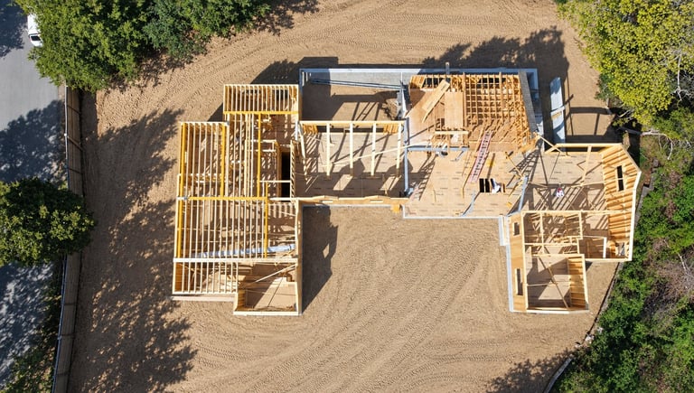 Aerial view of a residential construction site showing wooden framing and foundation layout on bare ground surrounded by trees.